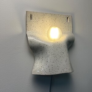 Może przedstawiać: Ścienna lampa ceramiczna o kremowej podstawie z czarnymi kropkami. Oprawa ma zakrzywiony, formowany kształt z dwoma prostokątnymi otworami montażowymi u g&oacute;ry oraz pojedynczą, świecącą, kulistą żar&oacute;wką umieszczoną centralnie na środku.