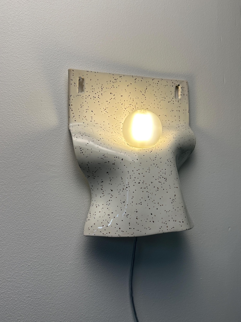 Może przedstawiać: Ścienna lampa ceramiczna o kremowej podstawie z czarnymi kropkami. Oprawa ma zakrzywiony, formowany kształt z dwoma prostokątnymi otworami montażowymi u g&oacute;ry oraz pojedynczą, świecącą, kulistą żar&oacute;wką umieszczoną centralnie na środku.