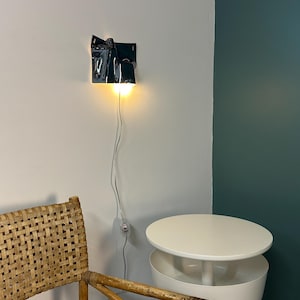 May include: A wall-mounted lamp with a glossy, dark, irregularly shaped, folded ceramic shade that emits a warm, downward-facing light. The lamp features a thin white power cord with an inline switch hanging vertically against a plain wall.