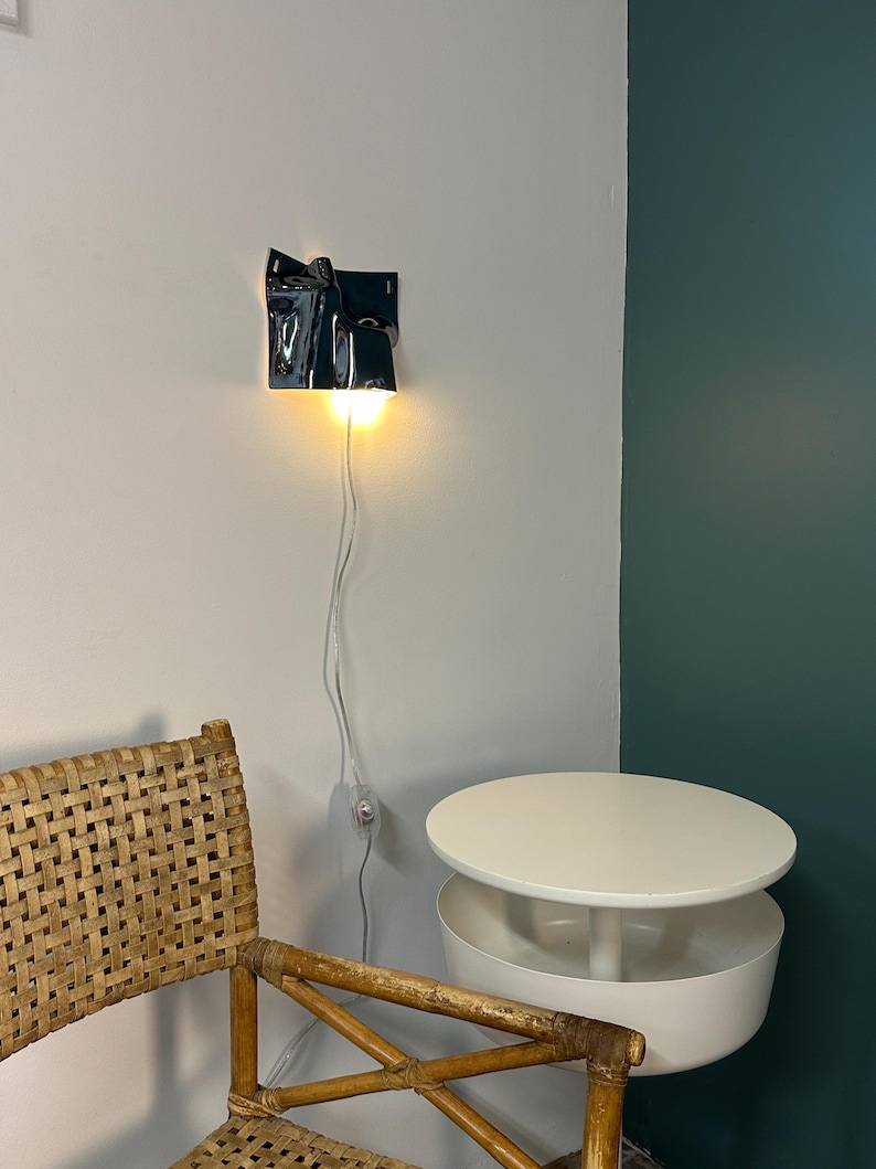 May include: A wall-mounted lamp with a glossy, dark, irregularly shaped, folded ceramic shade that emits a warm, downward-facing light. The lamp features a thin white power cord with an inline switch hanging vertically against a plain wall.