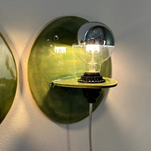May include: A wall-mounted light fixture featuring a circular, glossy green ceramic backplate with a small integrated shelf. A black socket holds a clear glass light bulb with a reflective silver-toned top half, emitting a warm glow.