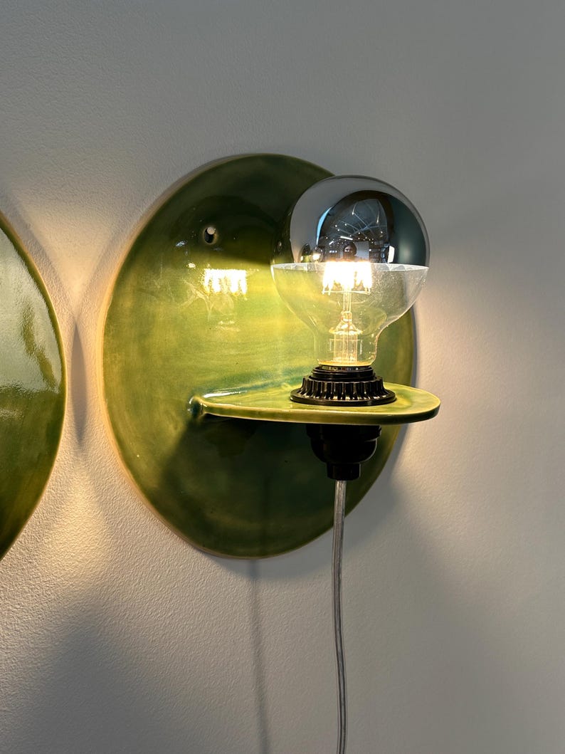 May include: A wall-mounted light fixture featuring a circular, glossy green ceramic backplate with a small integrated shelf. A black socket holds a clear glass light bulb with a reflective silver-toned top half, emitting a warm glow.
