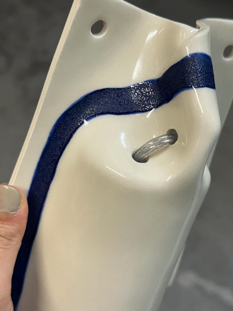 May include: A white ceramic wall-mounted object with a glossy finish and a textured, dark blue wavy line running vertically across the surface. The piece features two circular holes at the top and a central hole containing a clear plastic cord loop.
