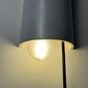 May include: A semi-cylindrical light gray ceramic wall sconce featuring a curved shade that directs light downward. A round, frosted glass light bulb is positioned beneath the shade, emitting a warm glow. A black power cord hangs vertically.