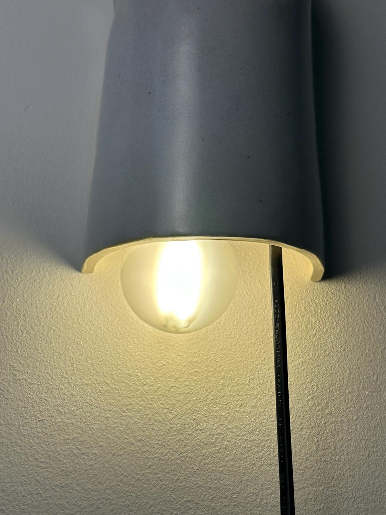 May include: A semi-cylindrical light gray ceramic wall sconce featuring a curved shade that directs light downward. A round, frosted glass light bulb is positioned beneath the shade, emitting a warm glow. A black power cord hangs vertically.