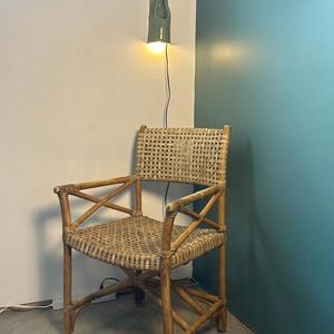 May include: A woven rattan armchair featuring a square backrest and seat with a cross-brace design on the armrests. The chair is constructed from light brown natural cane material and is positioned beneath a small, wall-mounted green ceramic lamp.