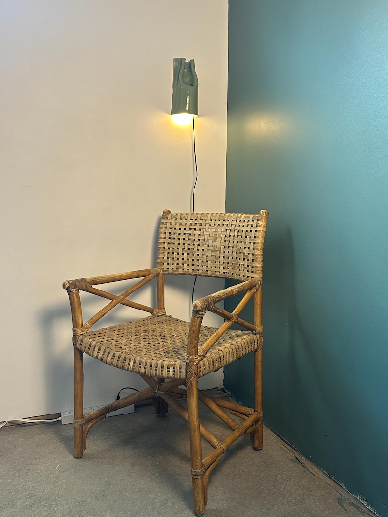 May include: A woven rattan armchair featuring a square backrest and seat with a cross-brace design on the armrests. The chair is constructed from light brown natural cane material and is positioned beneath a small, wall-mounted green ceramic lamp.