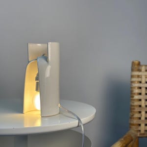 May include: A white ceramic table lamp with a curved, cylindrical structure that is open on one side. The lamp features a single exposed light bulb emitting a warm glow, connected to a clear power cord that extends from the base of the fixture.
