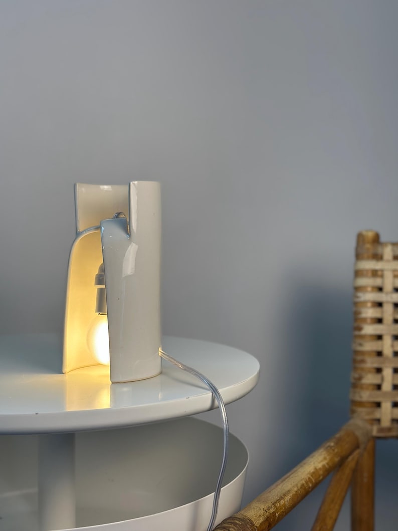 May include: A white ceramic table lamp with a curved, cylindrical structure that is open on one side. The lamp features a single exposed light bulb emitting a warm glow, connected to a clear power cord that extends from the base of the fixture.