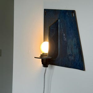 May include: A wall-mounted light fixture with a dark blue, geometric design. The fixture features a black bulb socket and a glowing, round light bulb. A gray cord extends from the base. The fixture is mounted on a white wall.