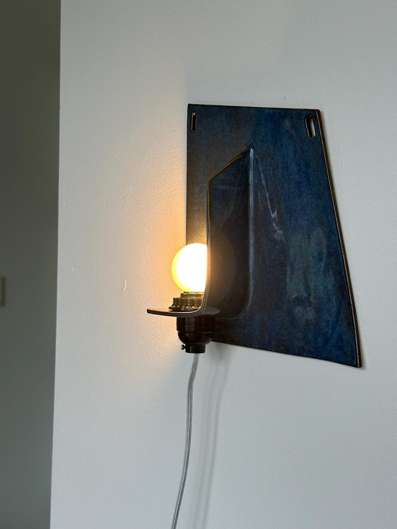 May include: A wall-mounted light fixture with a dark blue, geometric design. The fixture features a black bulb socket and a glowing, round light bulb. A gray cord extends from the base. The fixture is mounted on a white wall.