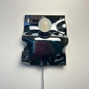 May include: A wall-mounted lamp featuring a glossy, dark, wavy ceramic backplate with a central black socket holding a round, frosted white light bulb. The ceramic piece has two small holes at the top for mounting and a thin power cord extending downward.