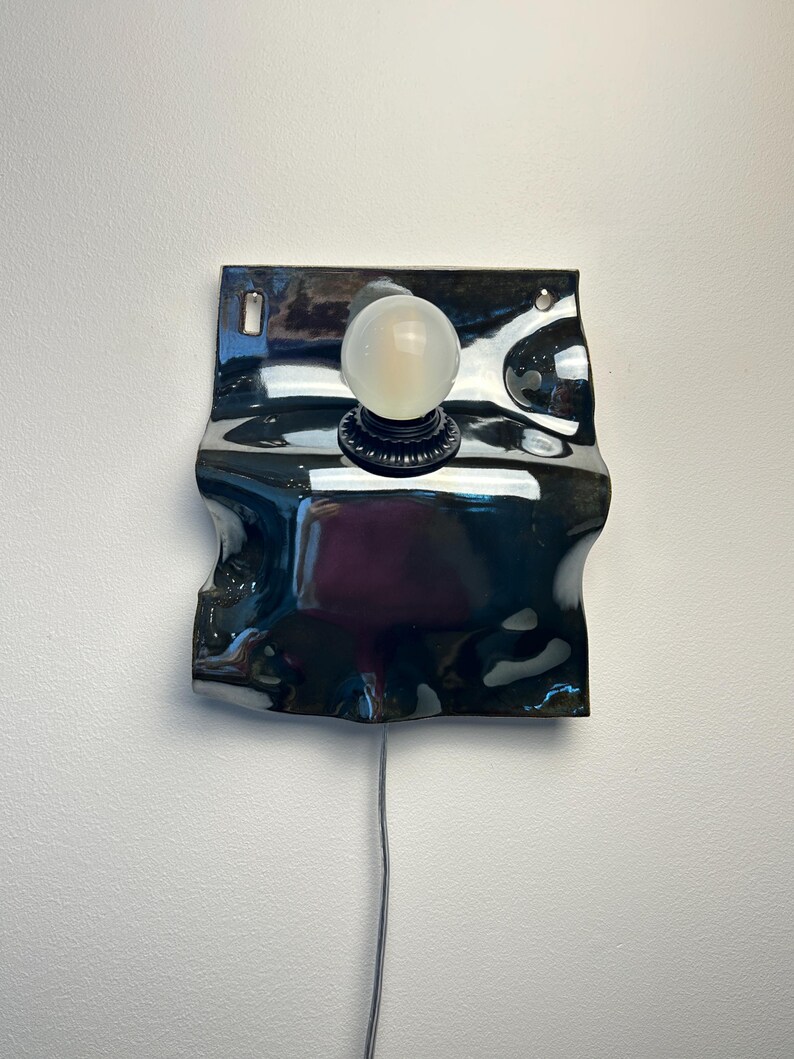 May include: A wall-mounted lamp featuring a glossy, dark, wavy ceramic backplate with a central black socket holding a round, frosted white light bulb. The ceramic piece has two small holes at the top for mounting and a thin power cord extending downward.