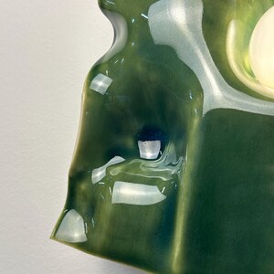 May include: A wall-mounted ceramic light fixture with a glossy, deep forest green glaze. The surface features an organic, undulating, and sculptural form with soft curves and indentations. A single, smooth, spherical white light bulb is visible.