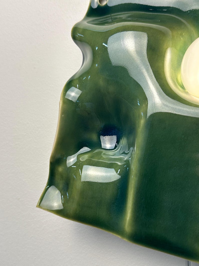 May include: A wall-mounted ceramic light fixture with a glossy, deep forest green glaze. The surface features an organic, undulating, and sculptural form with soft curves and indentations. A single, smooth, spherical white light bulb is visible.