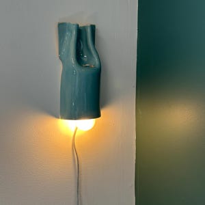 May include: A wall-mounted light fixture featuring a glossy, teal-colored ceramic shade with a fluid, organic, and elongated shape. The fixture emits a warm, glowing light from its base and includes a thin, light-colored electrical power cord.
