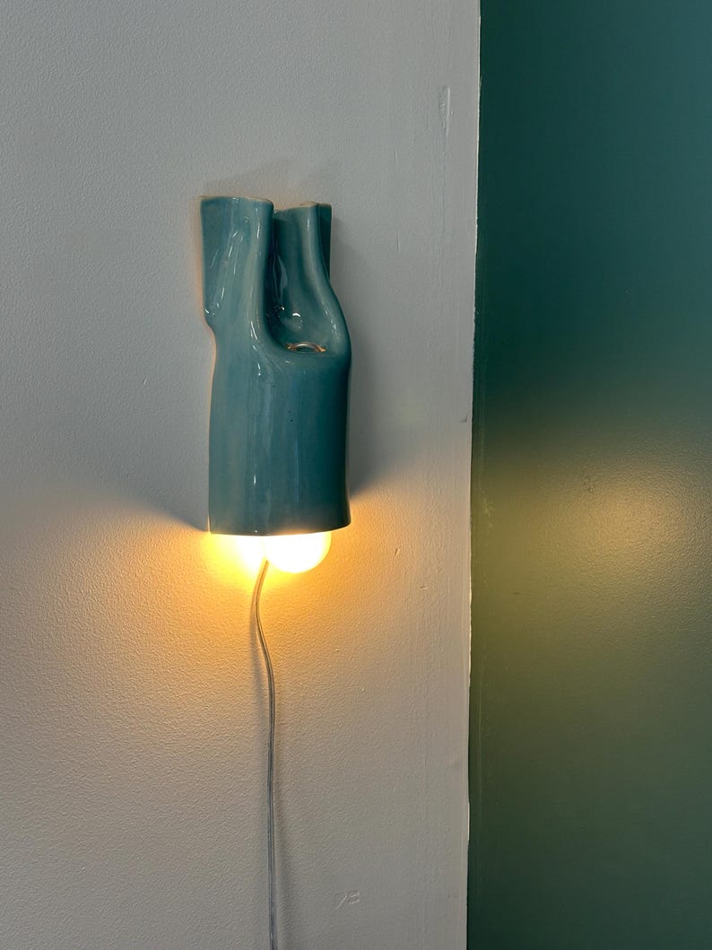 May include: A wall-mounted light fixture featuring a glossy, teal-colored ceramic shade with a fluid, organic, and elongated shape. The fixture emits a warm, glowing light from its base and includes a thin, light-colored electrical power cord.