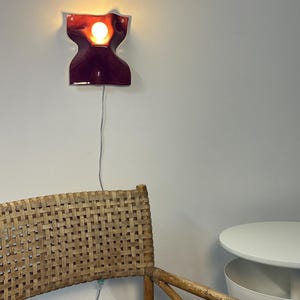 Puede incluir: Una l&aacute;mpara de pared con una pantalla de cer&aacute;mica brillante de color burdeos intenso en forma de reloj de arena. El aplique emite una luz c&aacute;lida desde una bombilla central y est&aacute; conectado a un cable el&eacute;ctrico fino y claro en la pared.