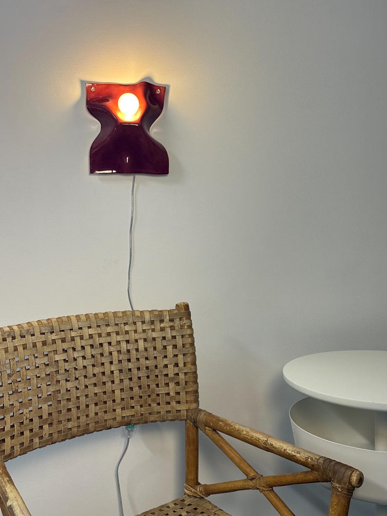 Puede incluir: Una l&aacute;mpara de pared con una pantalla de cer&aacute;mica brillante de color burdeos intenso en forma de reloj de arena. El aplique emite una luz c&aacute;lida desde una bombilla central y est&aacute; conectado a un cable el&eacute;ctrico fino y claro en la pared.