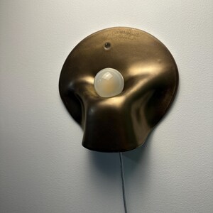 May include: A wall-mounted lamp featuring a circular, bronze-colored metallic shade with a soft, undulating, organic shape. A single, round, frosted white light bulb is positioned in the center of the shade, and a thin power cord extends downward.