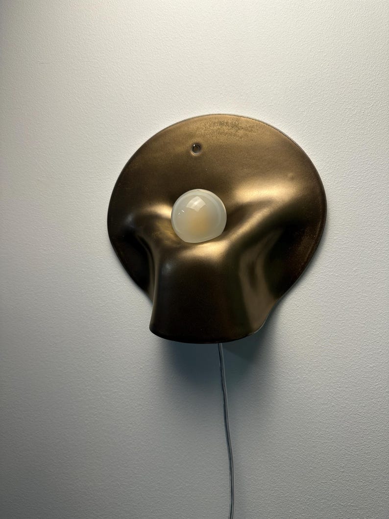 May include: A wall-mounted lamp featuring a circular, bronze-colored metallic shade with a soft, undulating, organic shape. A single, round, frosted white light bulb is positioned in the center of the shade, and a thin power cord extends downward.