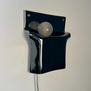 Peut inclure: Une applique murale en c&eacute;ramique noire brillante avec une plaque arri&egrave;re rectangulaire et une section avant incurv&eacute;e en forme de poche. Une ampoule ronde en verre d&eacute;poli blanc est log&eacute;e dans l'ouverture sup&eacute;rieure, aliment&eacute;e par un cordon blanc.