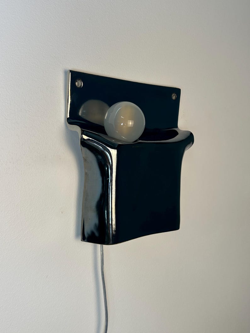 Peut inclure: Une applique murale en c&eacute;ramique noire brillante avec une plaque arri&egrave;re rectangulaire et une section avant incurv&eacute;e en forme de poche. Une ampoule ronde en verre d&eacute;poli blanc est log&eacute;e dans l'ouverture sup&eacute;rieure, aliment&eacute;e par un cordon blanc.