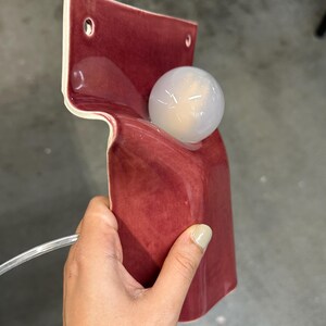 Pode incluir: Uma lumin&aacute;ria de parede em cer&acirc;mica com um acabamento vidrado em vermelho profundo e brilhante. A estrutura apresenta um painel traseiro plano com dois orif&iacute;cios de montagem circulares e uma sec&ccedil;&atilde;o frontal curva que segura uma l&acirc;mpada esf&eacute;rica.