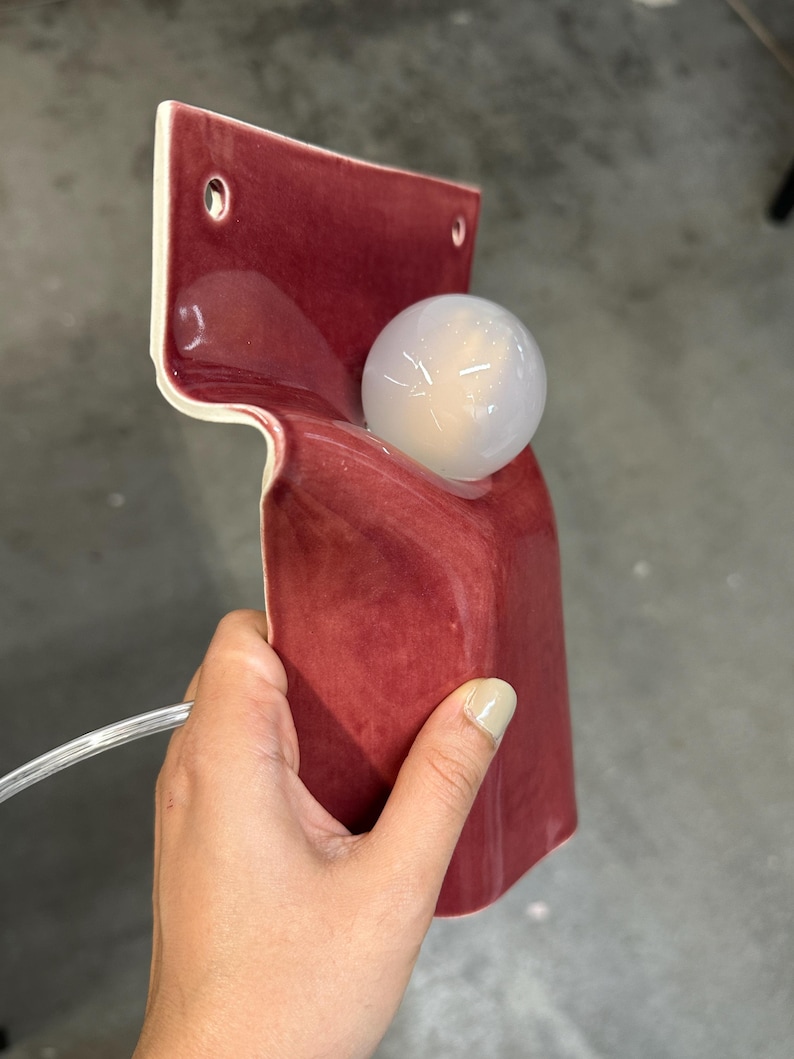 Pode incluir: Uma lumin&aacute;ria de parede em cer&acirc;mica com um acabamento vidrado em vermelho profundo e brilhante. A estrutura apresenta um painel traseiro plano com dois orif&iacute;cios de montagem circulares e uma sec&ccedil;&atilde;o frontal curva que segura uma l&acirc;mpada esf&eacute;rica.