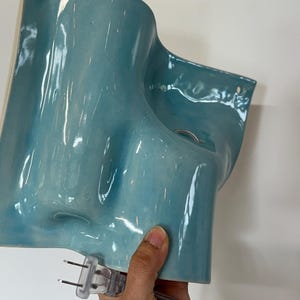 May include: A glossy, light blue ceramic wall sconce with an organic, undulating shape. The fixture features two circular mounting holes at the top corners and includes a clear electrical cord with a standard two-pin plug attached at the base.