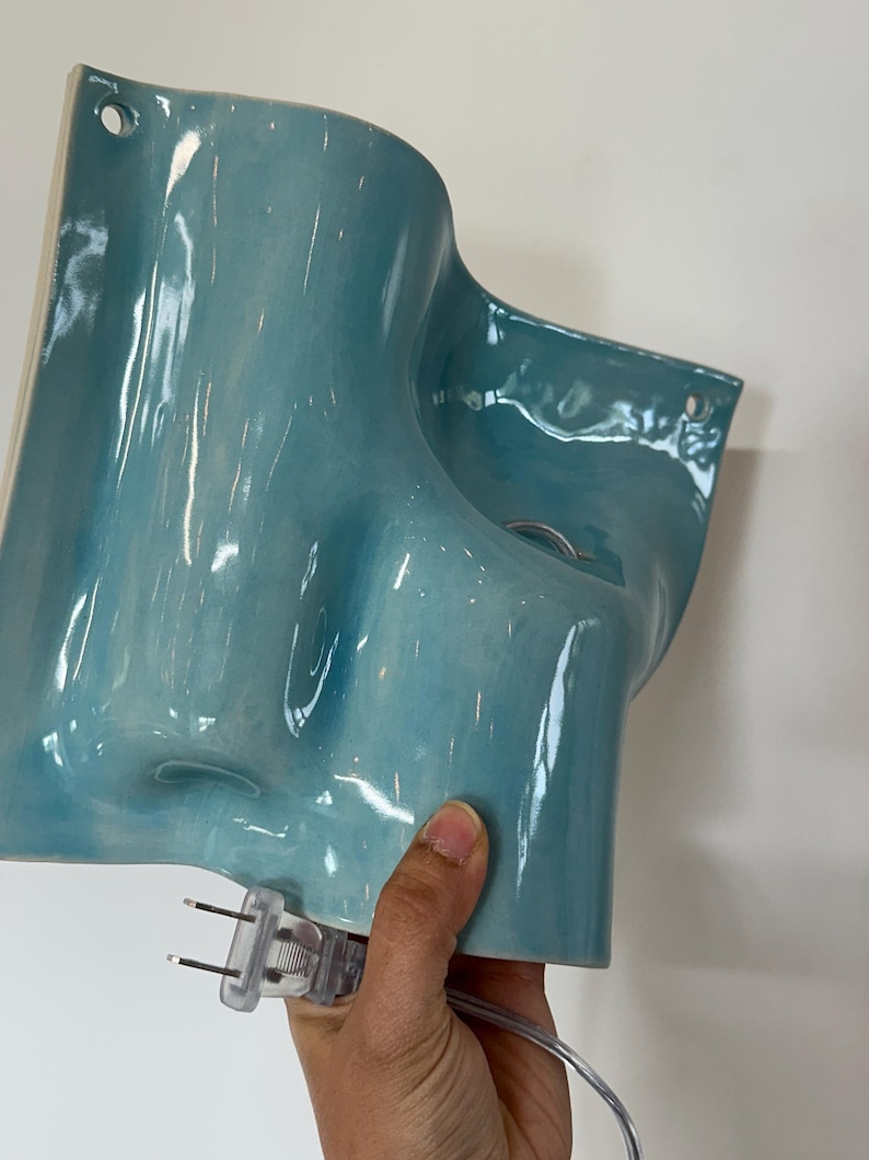 May include: A glossy, light blue ceramic wall sconce with an organic, undulating shape. The fixture features two circular mounting holes at the top corners and includes a clear electrical cord with a standard two-pin plug attached at the base.