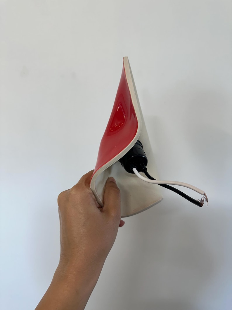 May include: A curved, cone-shaped lamp shade with a glossy red exterior and a matte white interior. A black plastic light socket is attached to the center, featuring two exposed electrical wires, one black and one white, extending from the base.