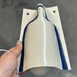 May include: A wall-mounted light fixture with a glossy off-white ceramic body shaped like a bottle. The fixture features a dark blue textured trim along the curved edges and two circular mounting holes at the top. A clear power cord is attached.