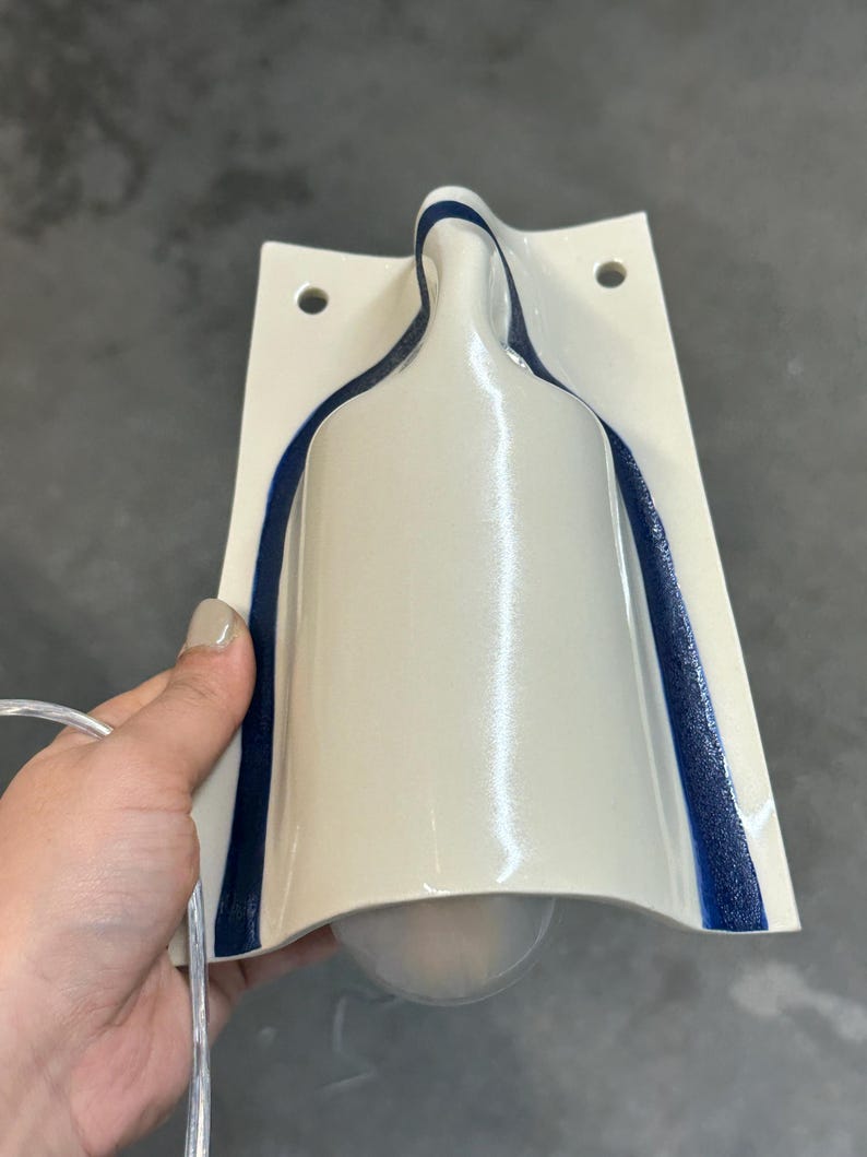 May include: A wall-mounted light fixture with a glossy off-white ceramic body shaped like a bottle. The fixture features a dark blue textured trim along the curved edges and two circular mounting holes at the top. A clear power cord is attached.