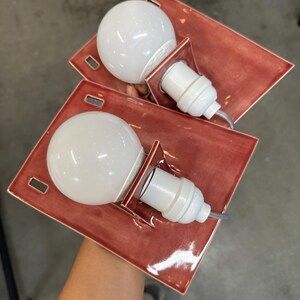 Puede incluir: Dos apliques de pared rectangulares con una base de cer&aacute;mica de color terracota brillante y acabado liso. Cada l&aacute;mpara incluye un casquillo blanco y una bombilla redonda grande de cristal blanco esmerilado en el centro.