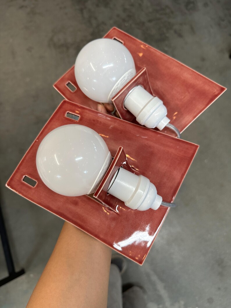 Puede incluir: Dos apliques de pared rectangulares con una base de cer&aacute;mica de color terracota brillante y acabado liso. Cada l&aacute;mpara incluye un casquillo blanco y una bombilla redonda grande de cristal blanco esmerilado en el centro.