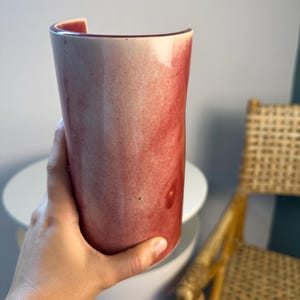 May include: A cylindrical ceramic vase featuring a smooth, glossy finish with a gradient of deep red and soft pink hues. The vessel has an open, curved top edge and a solid, rounded base, showcasing a blended, watercolor-like appearance.