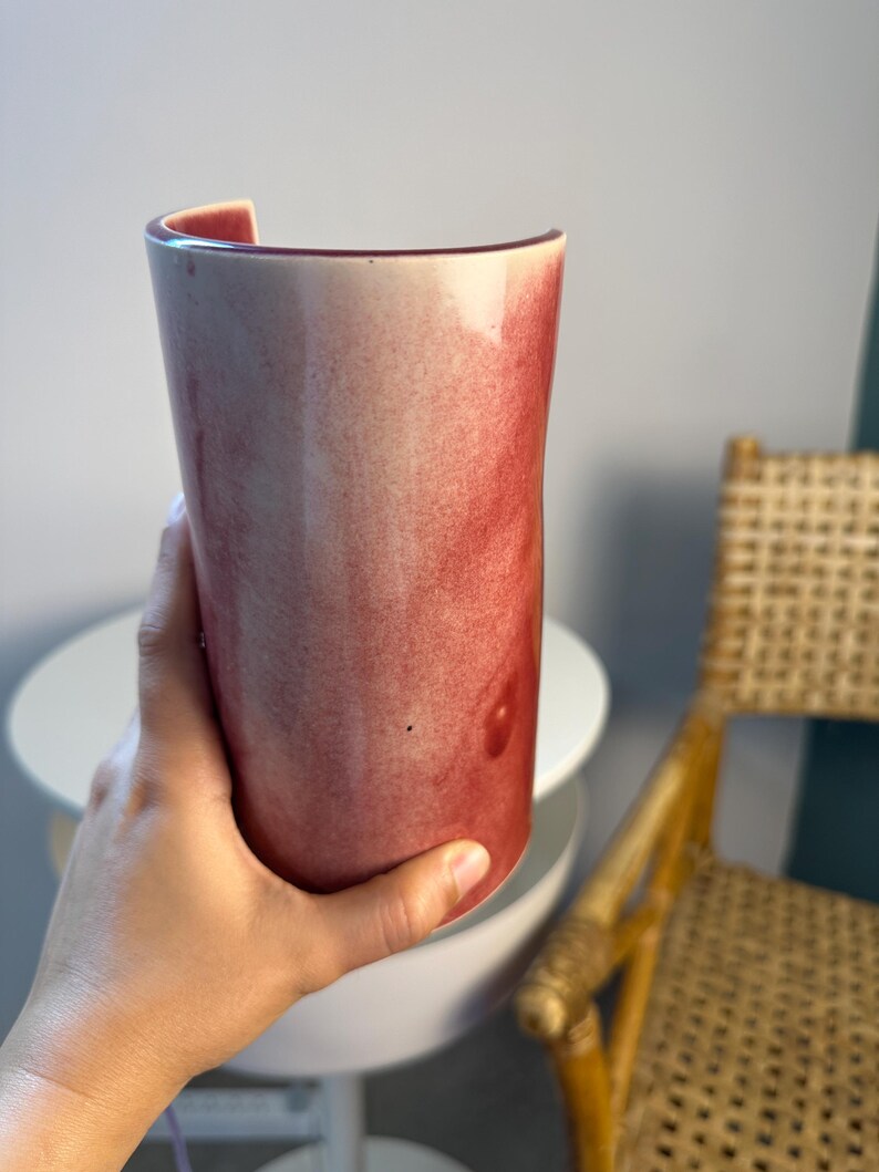 May include: A cylindrical ceramic vase featuring a smooth, glossy finish with a gradient of deep red and soft pink hues. The vessel has an open, curved top edge and a solid, rounded base, showcasing a blended, watercolor-like appearance.