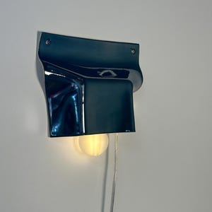 May include: A dark blue wall-mounted lamp with a glossy, curved ceramic shade. The fixture features a rectangular top section with two visible mounting screws and a downward-facing opening that emits a warm, soft glow from a round light bulb.