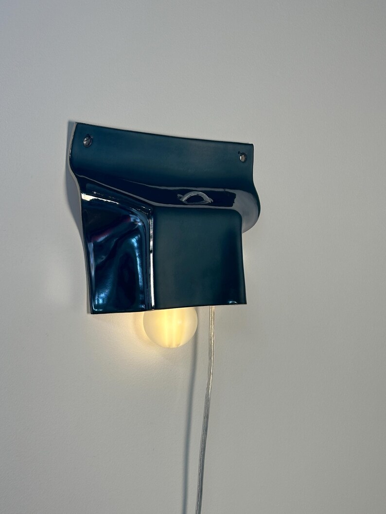 May include: A dark blue wall-mounted lamp with a glossy, curved ceramic shade. The fixture features a rectangular top section with two visible mounting screws and a downward-facing opening that emits a warm, soft glow from a round light bulb.
