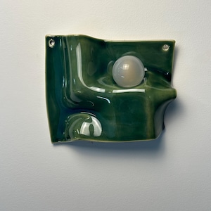 May include: A wall-mounted ceramic light fixture with a glossy, deep green, undulating surface. The piece features a smooth, spherical, translucent white glass bulb integrated into the center of the wave-like form, with two small metal mounting holes.