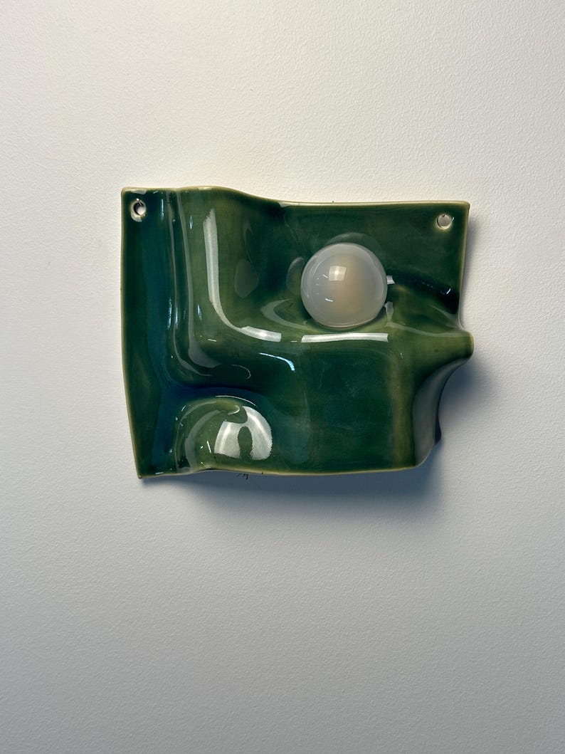 May include: A wall-mounted ceramic light fixture with a glossy, deep green, undulating surface. The piece features a smooth, spherical, translucent white glass bulb integrated into the center of the wave-like form, with two small metal mounting holes.
