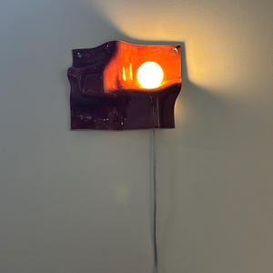 May include: A wall-mounted lamp featuring a deep purple, wavy, ceramic-like rectangular shade with a glossy finish. A single round, warm-toned light bulb is positioned in the upper right section, casting a soft glow against the dark surface.
