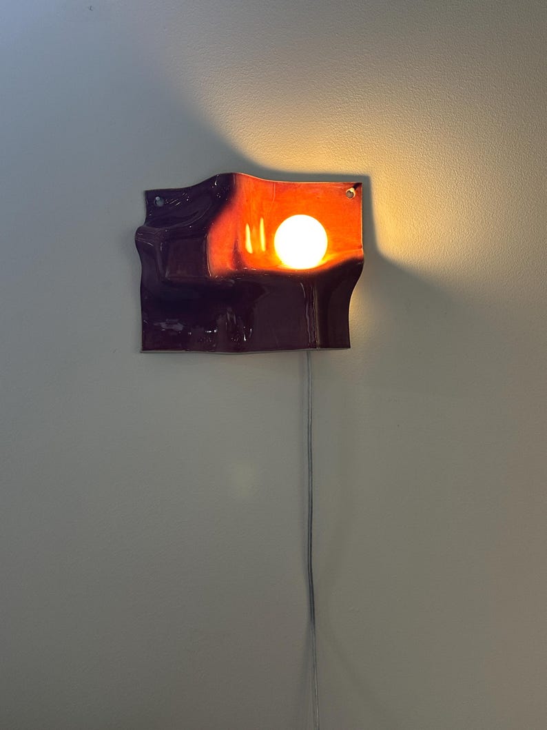 May include: A wall-mounted lamp featuring a deep purple, wavy, ceramic-like rectangular shade with a glossy finish. A single round, warm-toned light bulb is positioned in the upper right section, casting a soft glow against the dark surface.