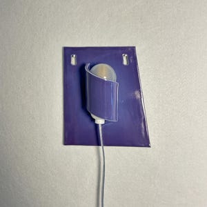 Pode incluir: Uma arandela de parede moderna com uma placa traseira retangular roxa e uma lumin&aacute;ria curva e cil&iacute;ndrica. A lumin&aacute;ria cont&eacute;m uma l&acirc;mpada redonda fosca e est&aacute; conectada a um cabo branco fino. A arandela &eacute; montada em uma parede cinza claro.