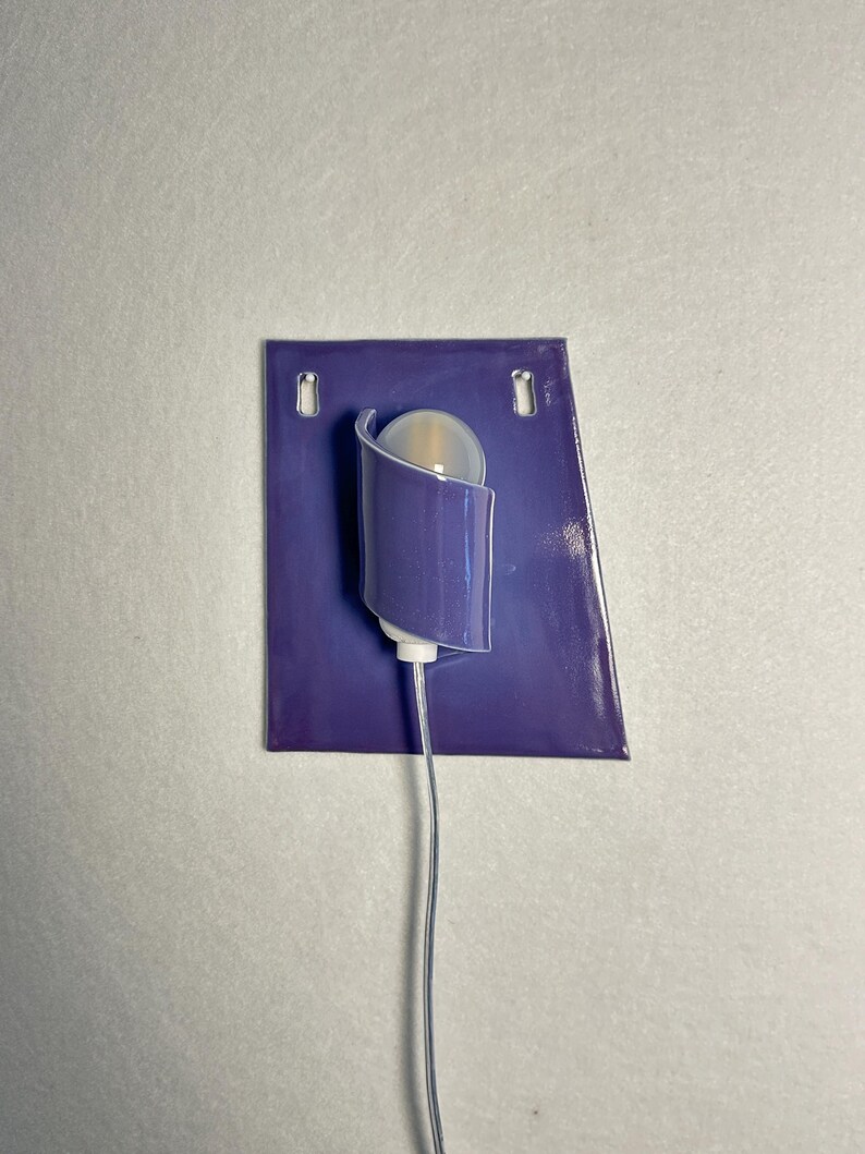 Pode incluir: Uma arandela de parede moderna com uma placa traseira retangular roxa e uma lumin&aacute;ria curva e cil&iacute;ndrica. A lumin&aacute;ria cont&eacute;m uma l&acirc;mpada redonda fosca e est&aacute; conectada a um cabo branco fino. A arandela &eacute; montada em uma parede cinza claro.