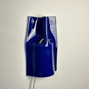 May include: A glossy, deep blue ceramic wall sconce with a sculptural, undulating form. The fixture features two small circular mounting holes at the top and a central cylindrical opening that houses a light socket with a visible silver cord.