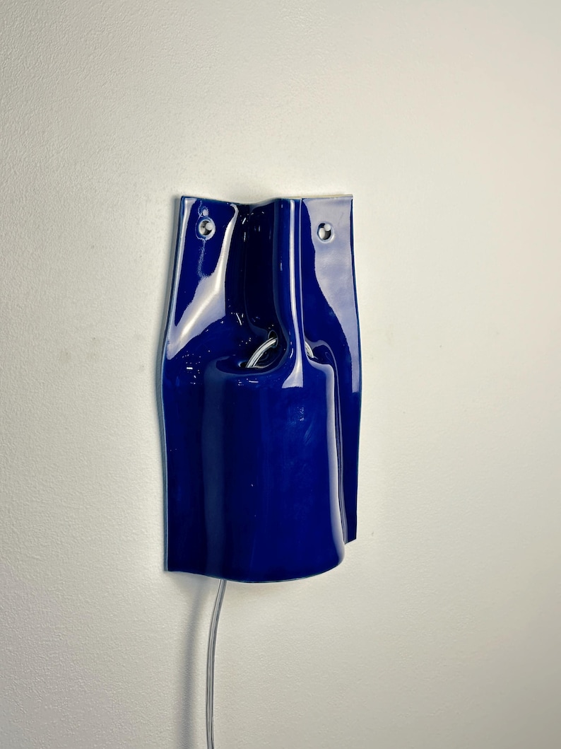 Puede incluir: Un aplique de pared de cer&aacute;mica en color azul profundo brillante con una forma escultural y ondulada. El accesorio cuenta con dos peque&ntilde;os orificios de montaje circulares en la parte superior y una abertura cil&iacute;ndrica central que alberga un casquillo con un cable plateado visible.