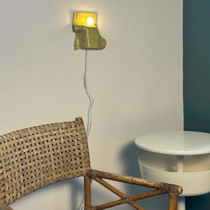Puede incluir: Un aplique de pared de cer&aacute;mica con un acabado brillante en color verde claro moteado. La l&aacute;mpara presenta una forma org&aacute;nica curva y emite una luz c&aacute;lida desde una bombilla. Un cable el&eacute;ctrico blanco y fino cuelga verticalmente hacia abajo.