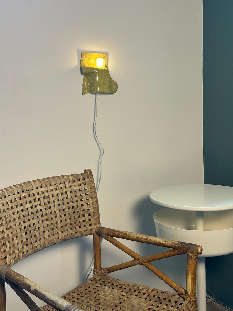 Puede incluir: Un aplique de pared de cer&aacute;mica con un acabado brillante en color verde claro moteado. La l&aacute;mpara presenta una forma org&aacute;nica curva y emite una luz c&aacute;lida desde una bombilla. Un cable el&eacute;ctrico blanco y fino cuelga verticalmente hacia abajo.
